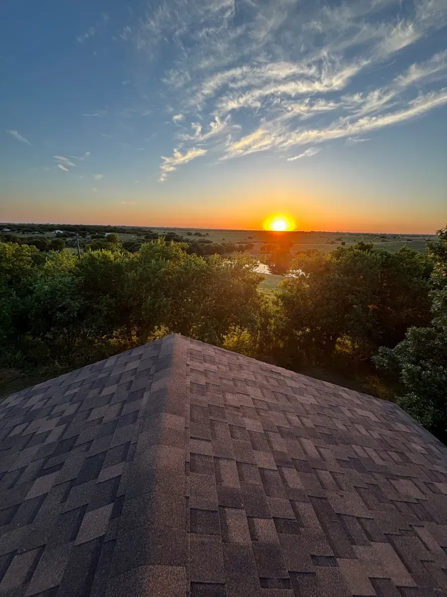 A sunset glow above a lake and trees, viewed from the peak of a textured, shingled roof.