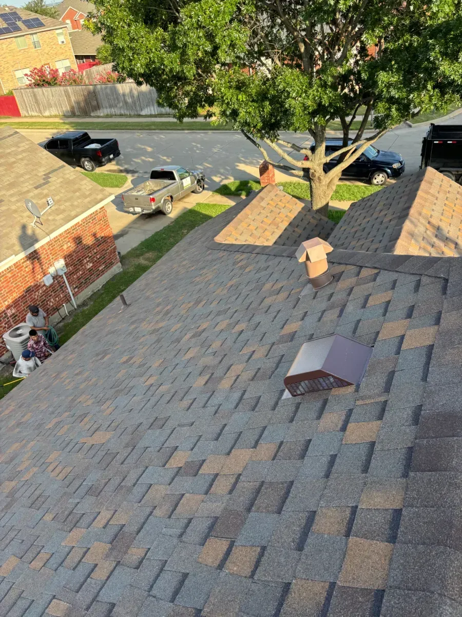 A high-angle view of a shingled roof with two vents, overlooking a residential street with cars and a tree.