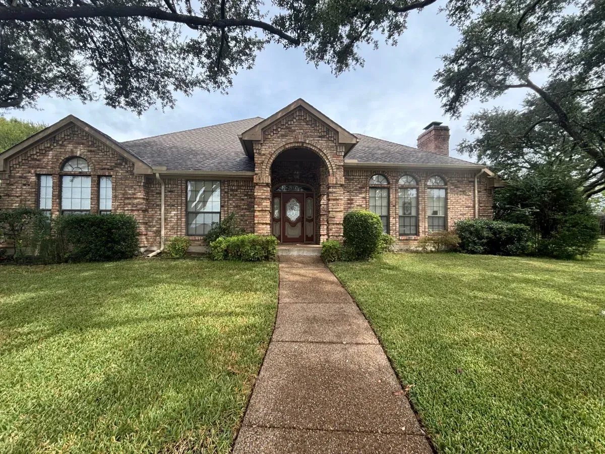 A one-story brick house with a arched entryway, central walkway, and large green lawn under trees.