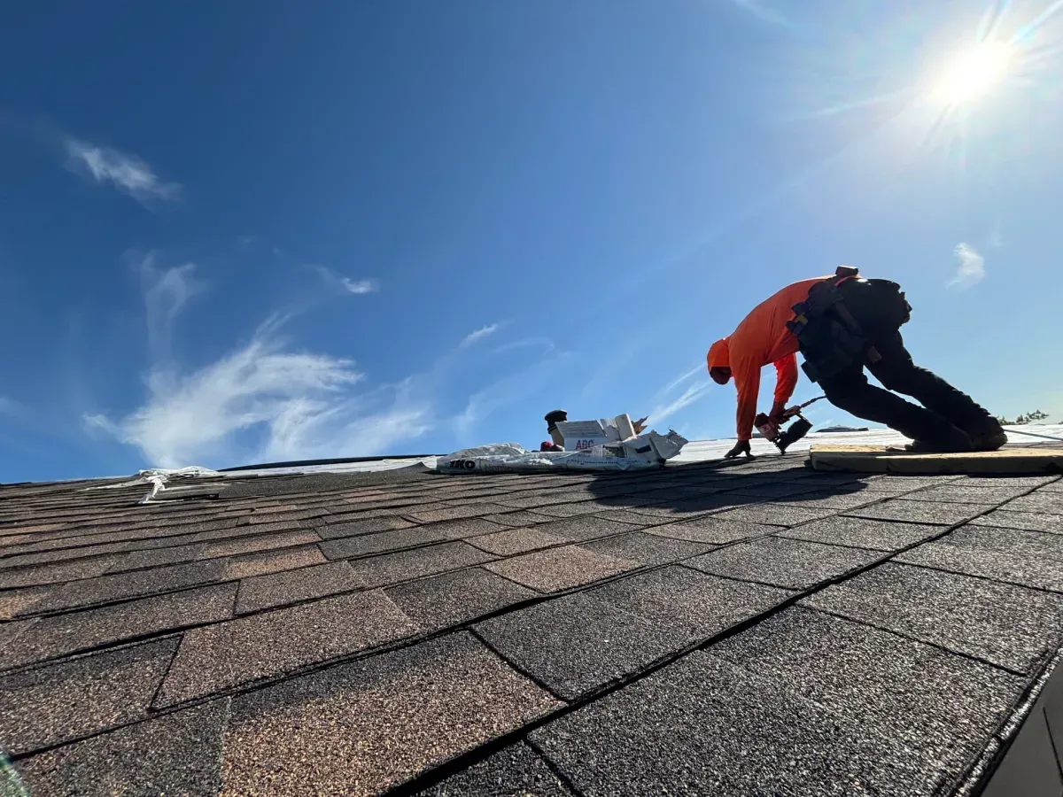 A worker in an orange hoodie using a nail gun to install dark asphalt shingles on a sloped roof under a clear blue sky.