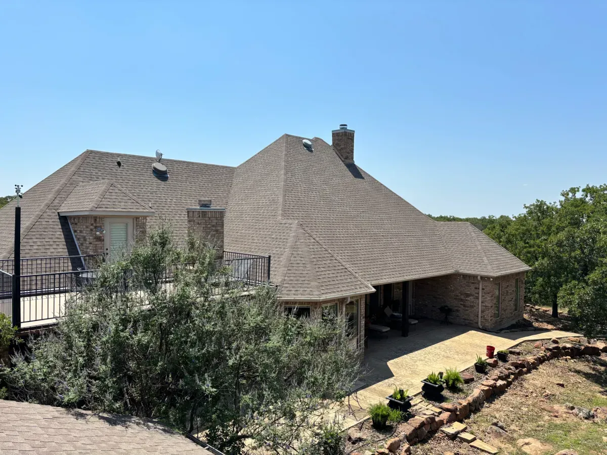 A tan brick house with a complex shingled roof, a stone patio, and a metal deck railing set against a wooded landscape.