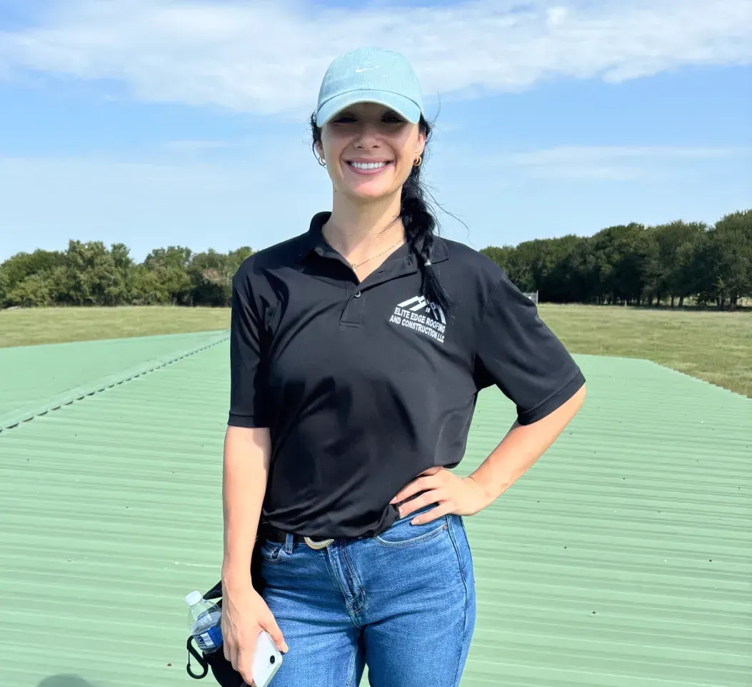 A smiling person in a black polo, blue jeans, and a light blue cap standing on a green roof outdoors.