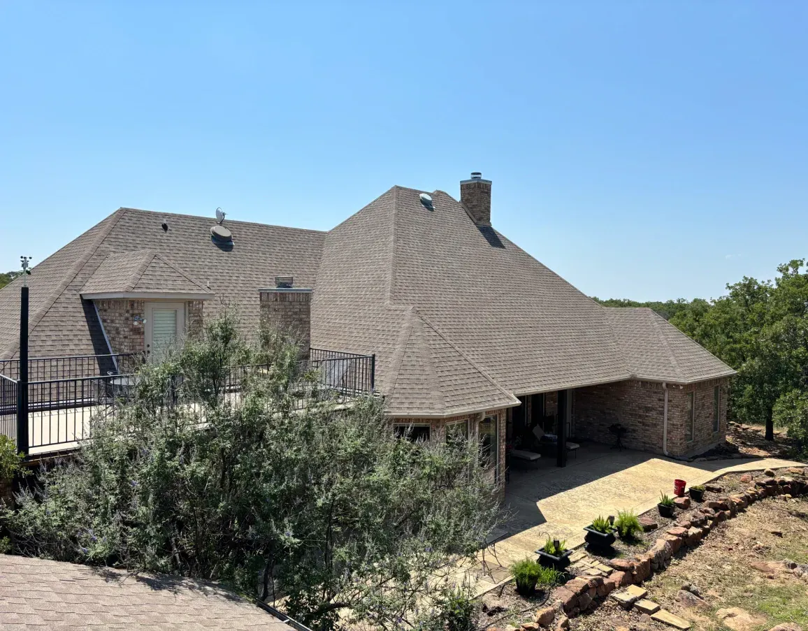A stone house with an intricate shingled roof features a terrace, porch, and landscaped garden under a clear blue sky.