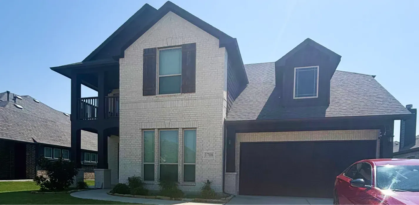 A two-story house with cream-colored brick, dark wood trim, a front balcony, a dormer window, and a red car parked outside.