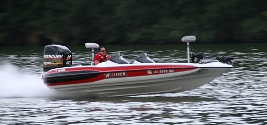 A red and white motorboat speeds across the water. A person is driving the boat. Trees line the background.