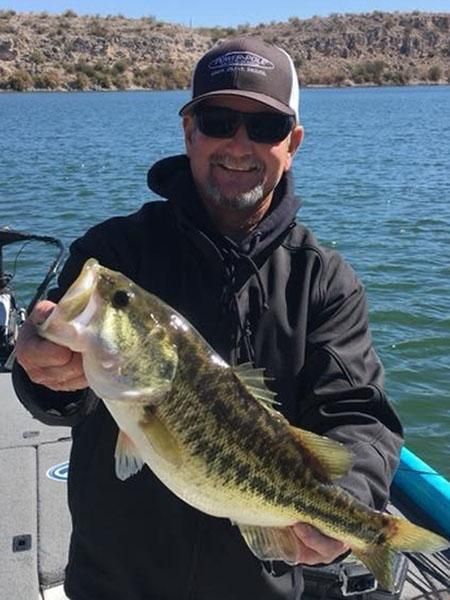 Man holding a largemouth bass on a boat, smiling, with a lake and hills in the background.