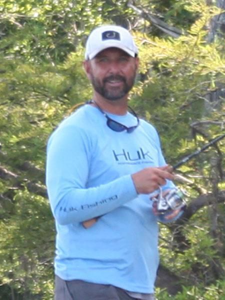 Man in blue shirt and white hat holding fishing rod near green foliage.