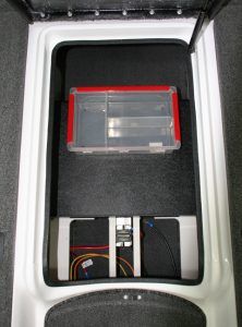 Storage compartment in a white boat, open. Red and clear tackle box on top. Lower section contains a device with wires.