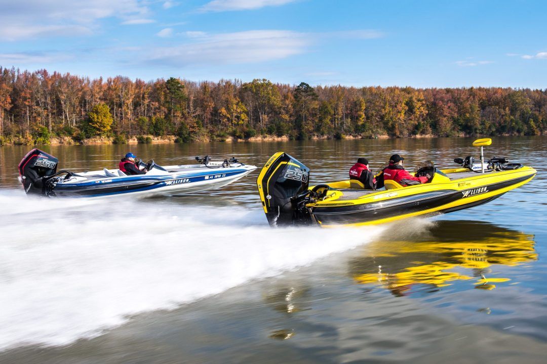 Two yellow and blue speedboats race across a lake. Fall foliage in the background.