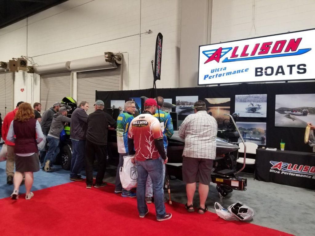People at an Allison Boats booth at an indoor event, red carpet in front, display with photos.