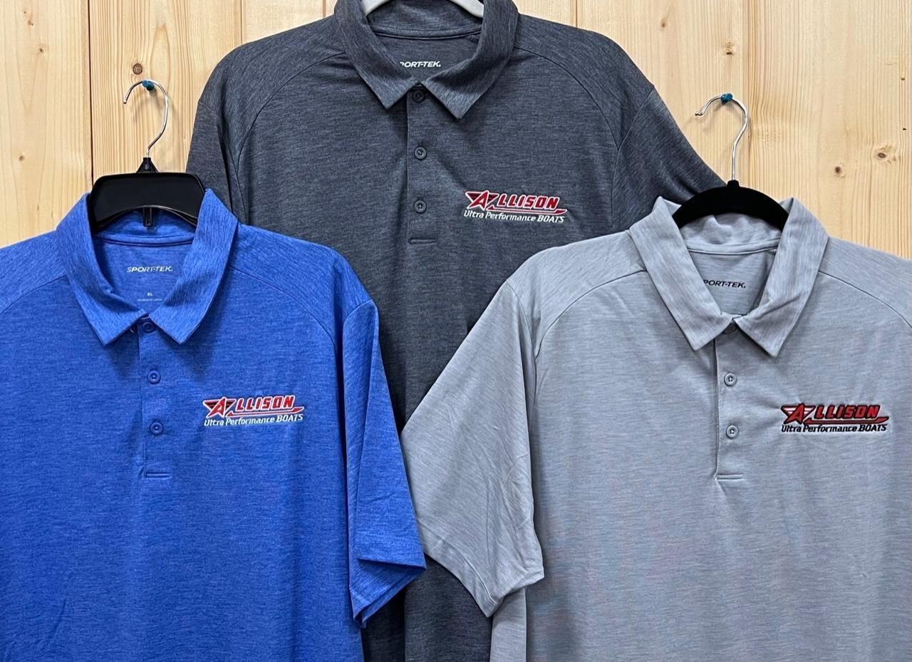 Three polo shirts in blue, gray, and light gray hanging on a wooden wall; each with a logo on the chest.