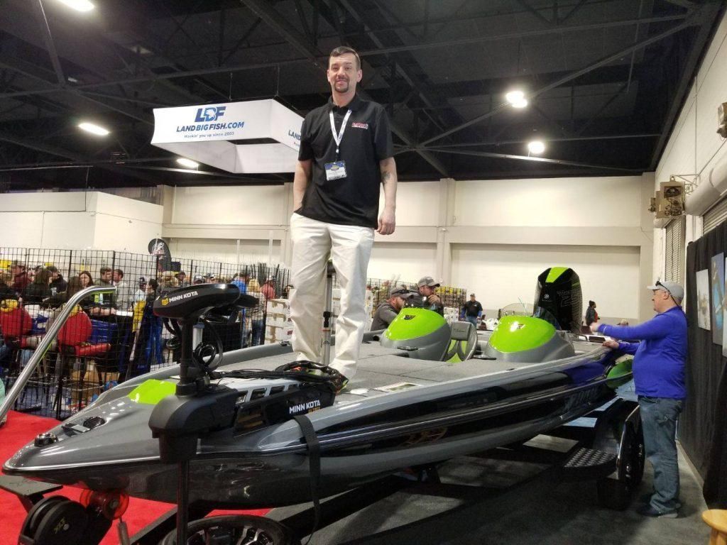Man standing on a bass boat display at a boat show. Others browse the exhibit.