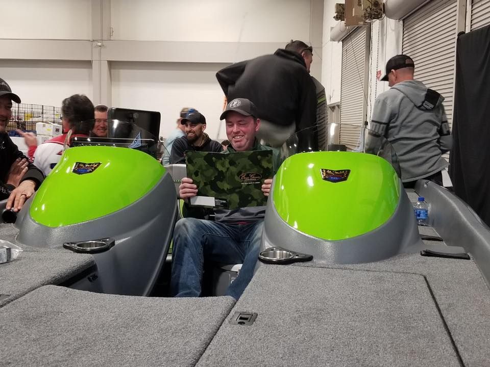 Man holding a camo design sits in a boat with green seats, surrounded by other men in a garage.