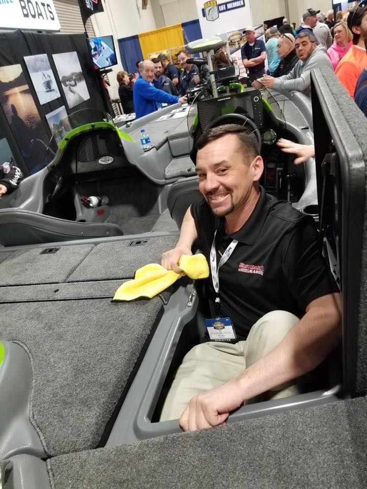 Man smiling in a boat, holding a yellow cloth, at a boat show. People in background.