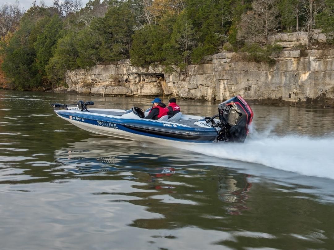 A blue and white bass boat speeds across water, two people on board, rocky shoreline background.