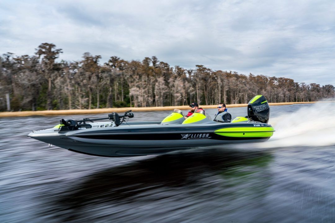 Bass boat speeding on water, neon green accents, two people onboard, trees in background.