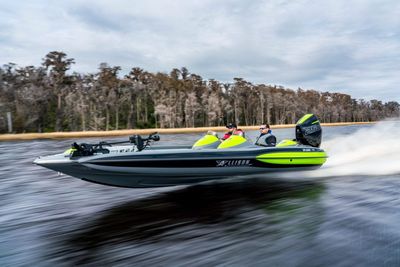 Bass boat speeding on water, neon green accents, two people onboard, trees in background.