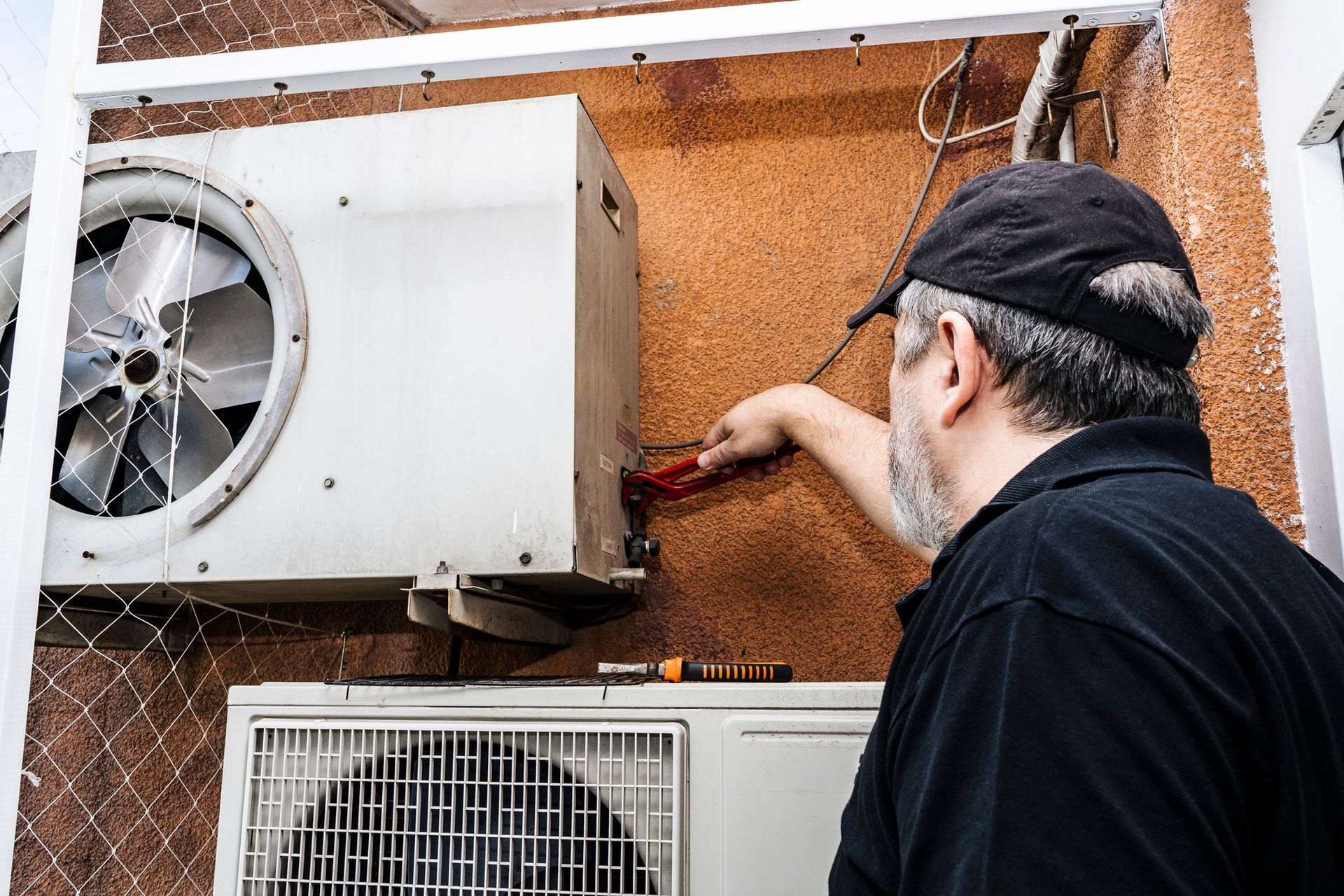 Worker adjusts wall-mounted AC unit with red-handled tool outdoors.