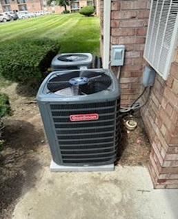 Air conditioning unit, Goodman brand, next to brick wall with electrical boxes, outdoors.