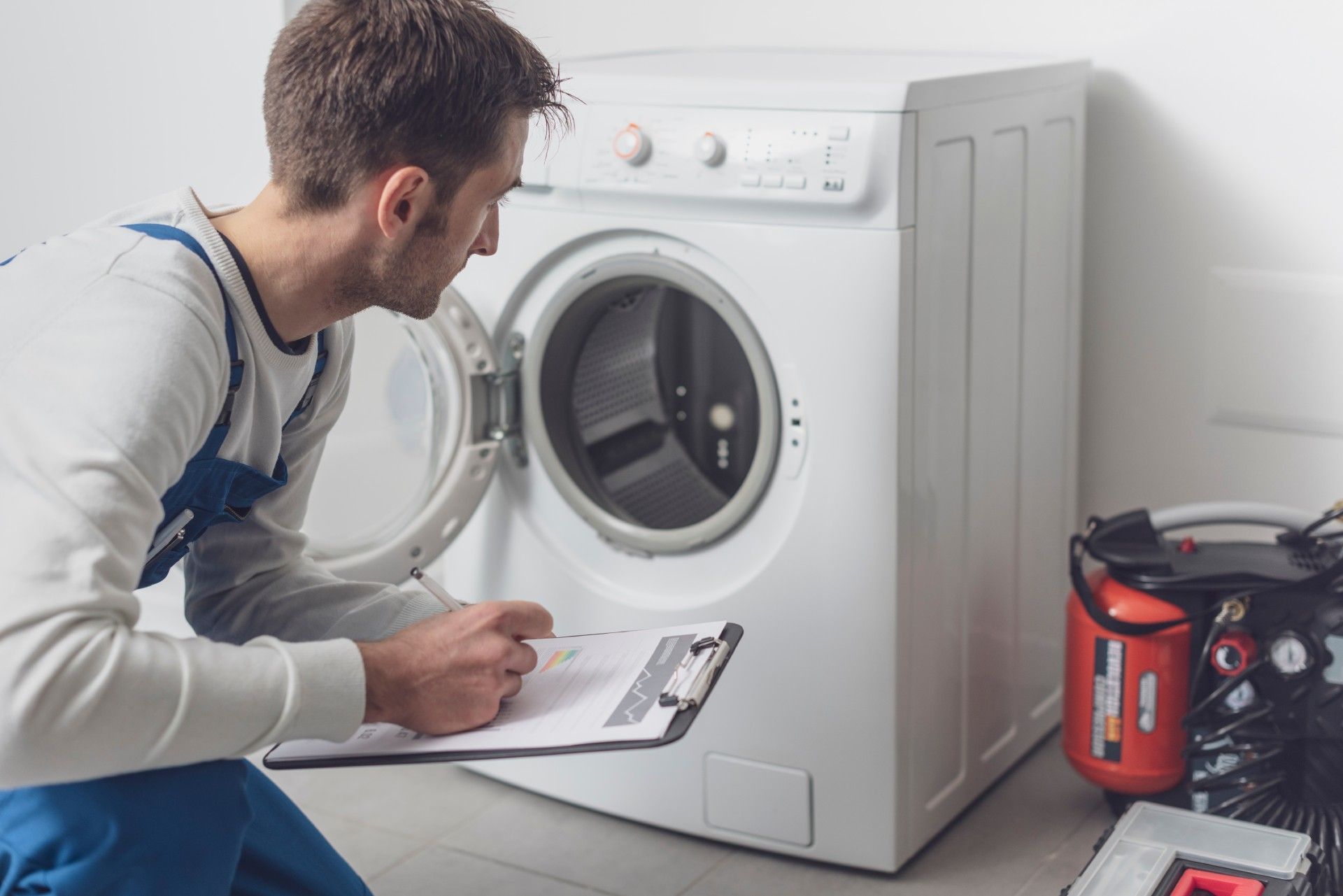 A man is kneeling down in front of a washing machine and writing on a clipboard.