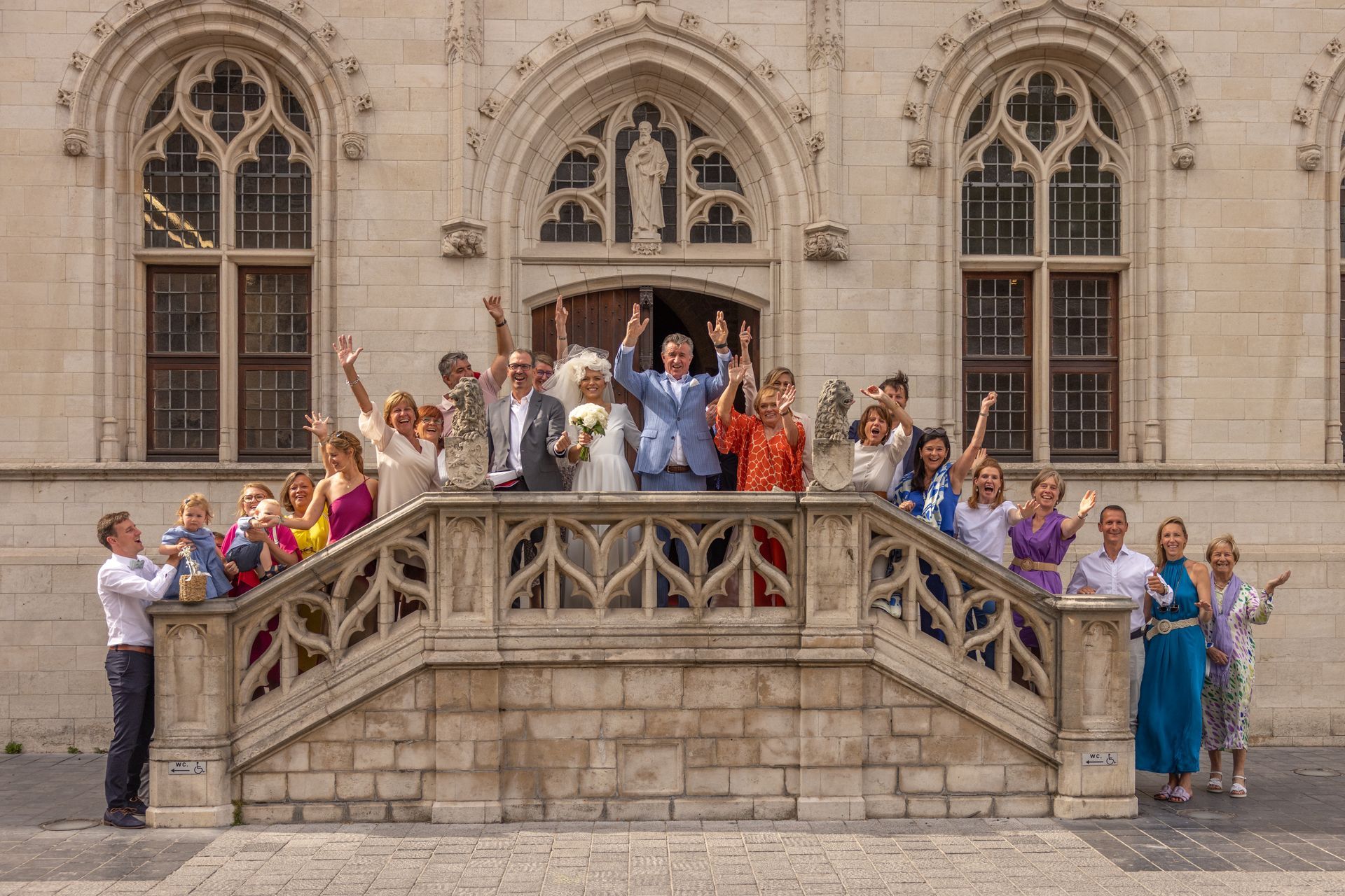 Een bruidsgezelschap op de stenen trap voor een sierlijk gebouw; mensen juichen met geheven armen.