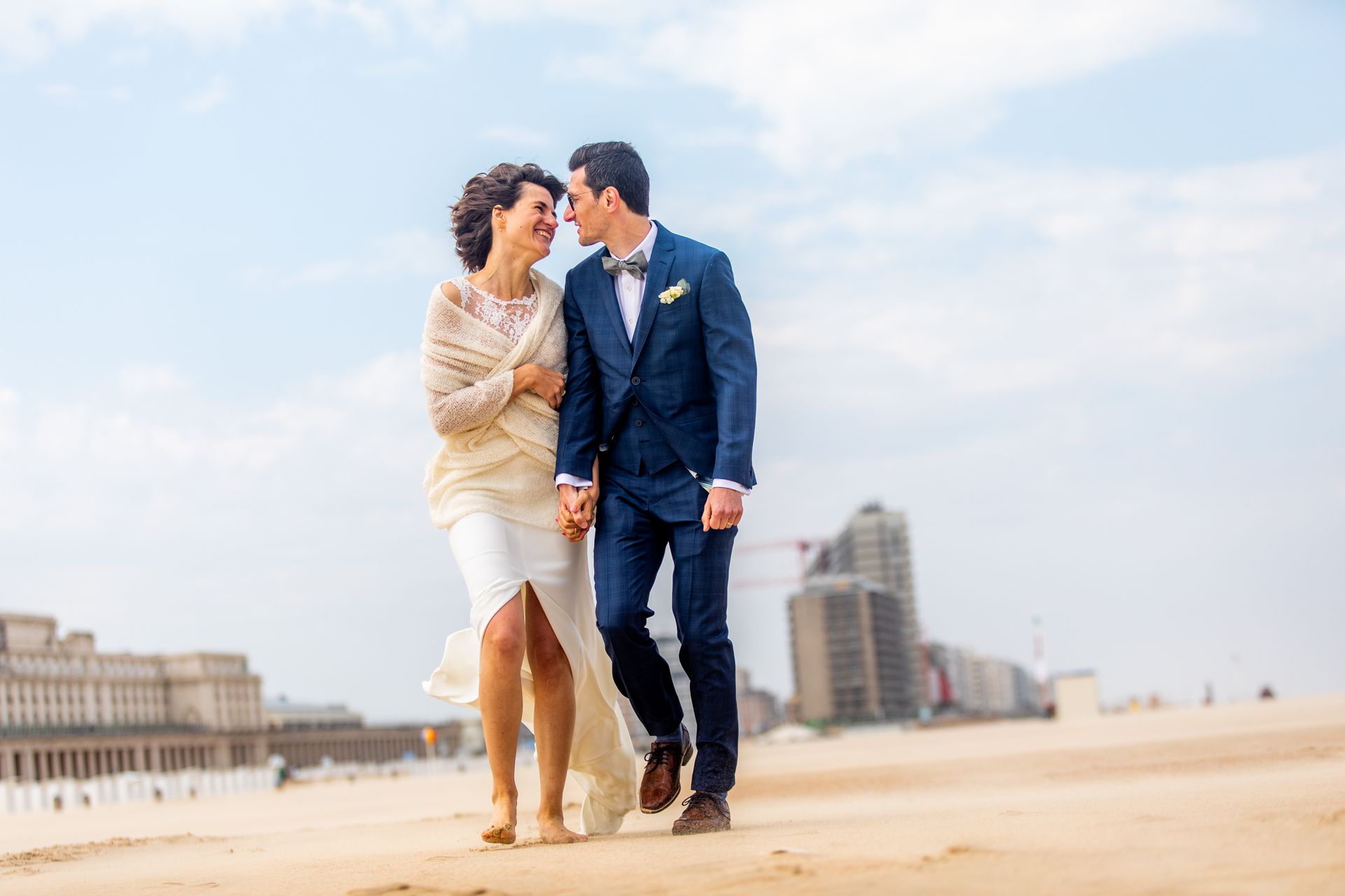 Een bruidspaar loopt lachend en hand in hand over het strand, met de gebouwen van de stad op de achtergrond.