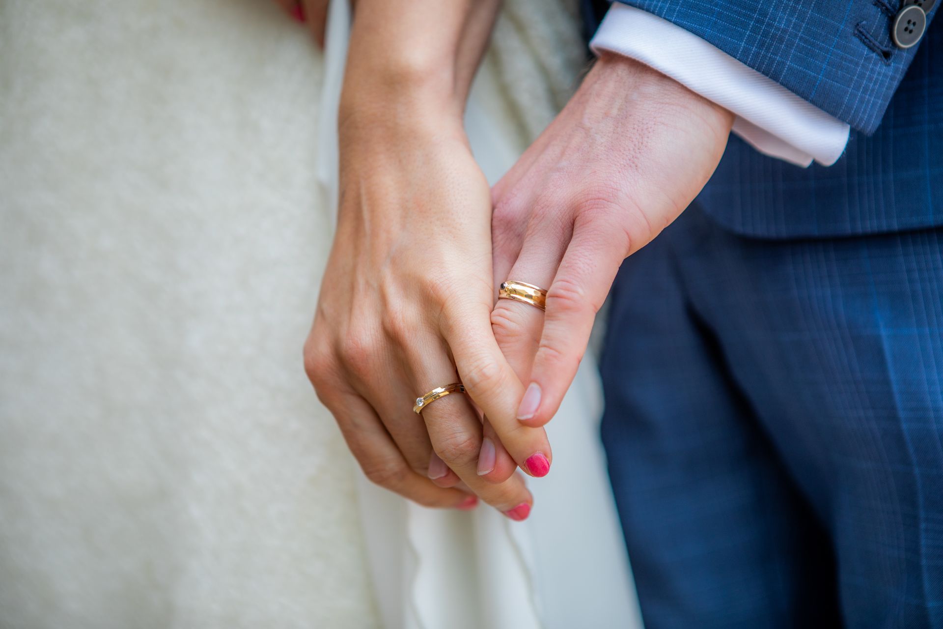 Trouwringen om handen, een vrouw in een witte jurk en een man in een blauw pak houden elkaars handen vast.