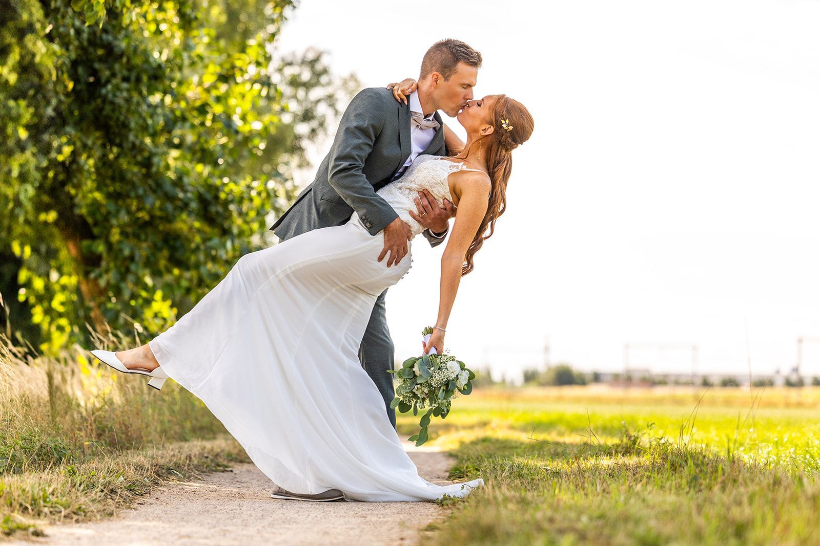Groom dips bride for a kiss on a dirt path in a sunny field. She wears a white gown, he wears a gray suit.