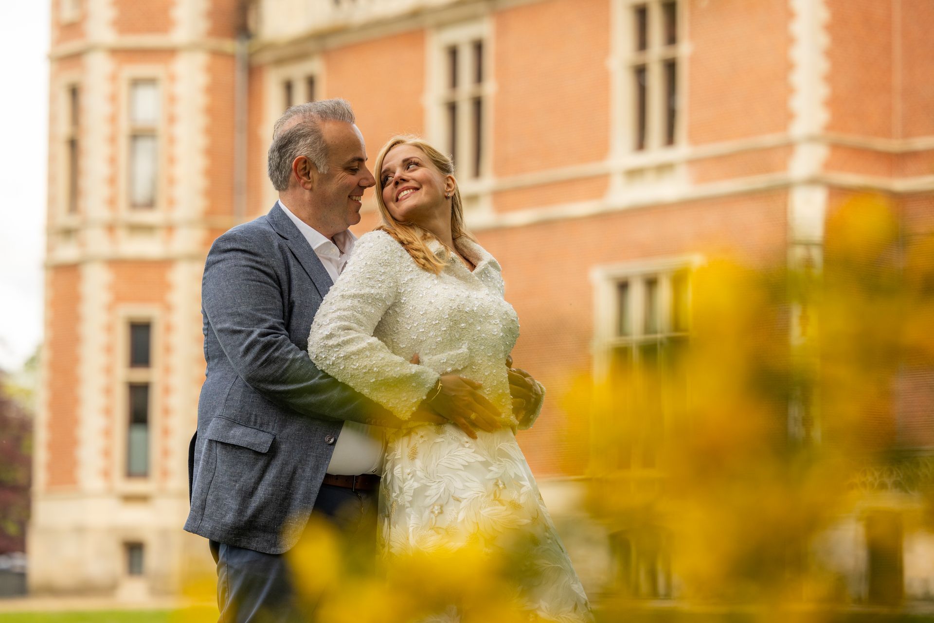 Een stel omhelst elkaar en lacht naar elkaar voor een rood bakstenen gebouw met gele bloemen op de voorgrond.