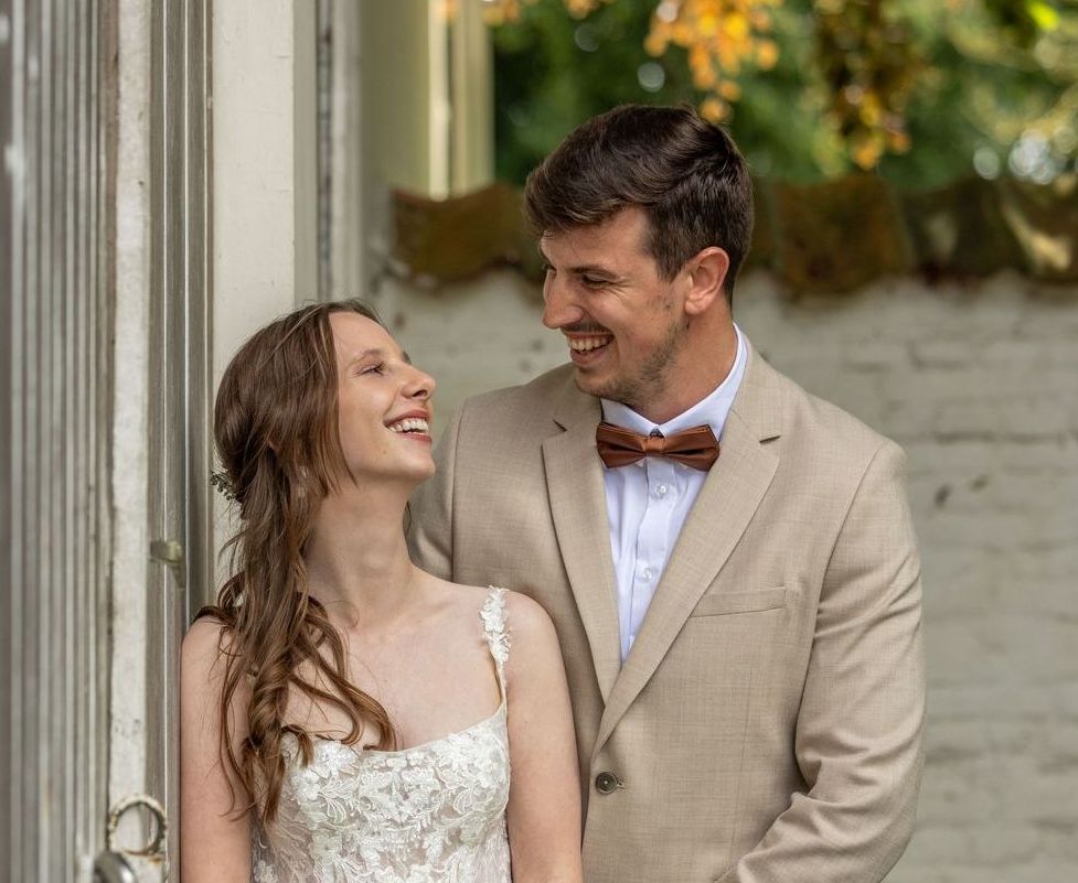 Bruid en bruidegom lachen naar elkaar; man in bruin pak met houten vlinderdas; vrouw in witte kanten jurk.