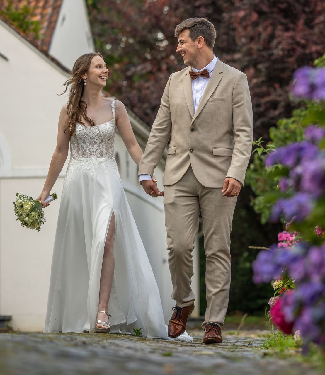 Bruid en bruidegom lopen hand in hand. Bruid in witte jurk, bruidegom in bruin pak. Paarse bloemen omlijsten het pad.