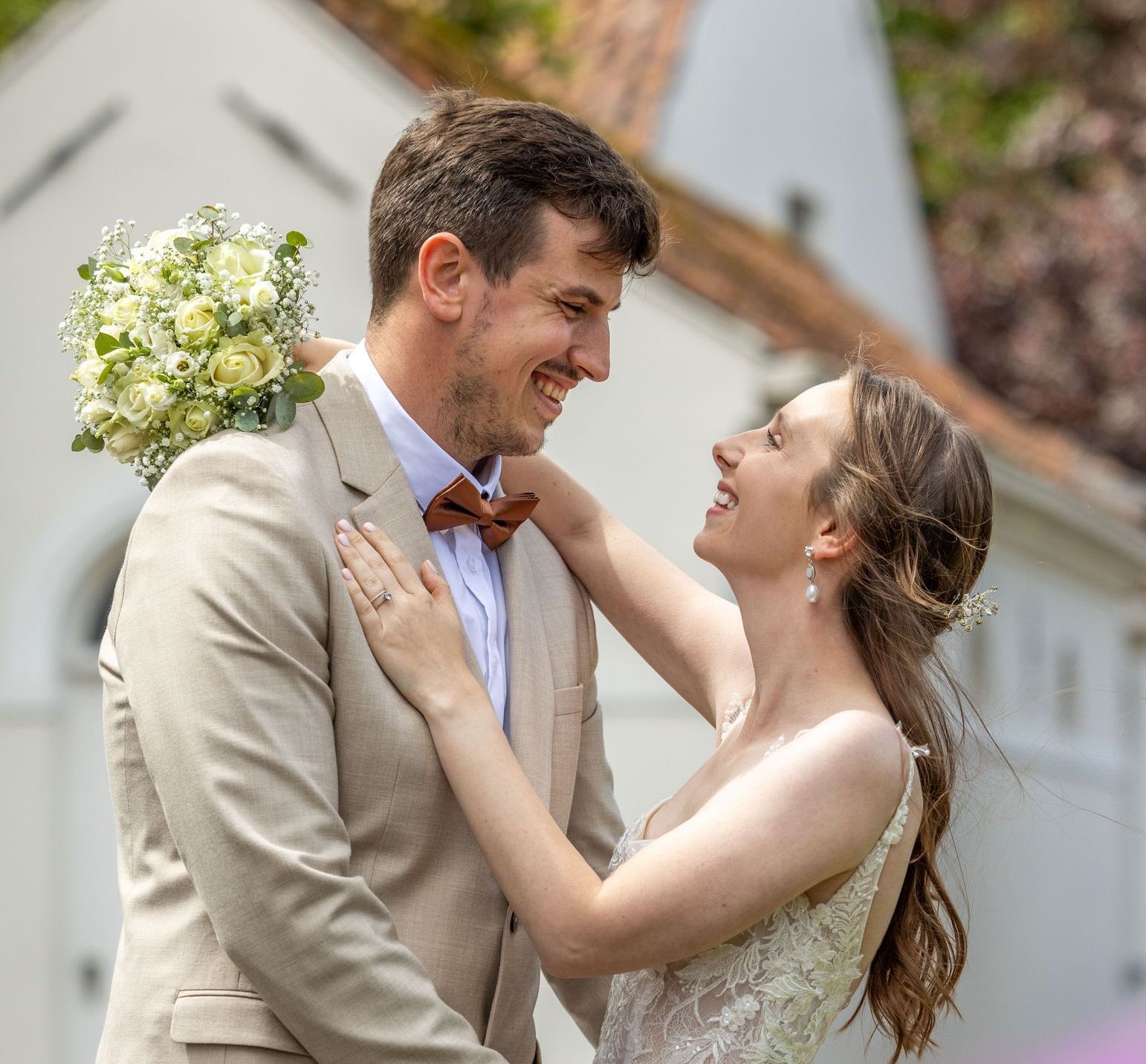 Het pasgetrouwde stel omhelst elkaar en lacht; de man in het beige pak, de vrouw in de witte jurk, op de achtergrond een klein wit kerkje.