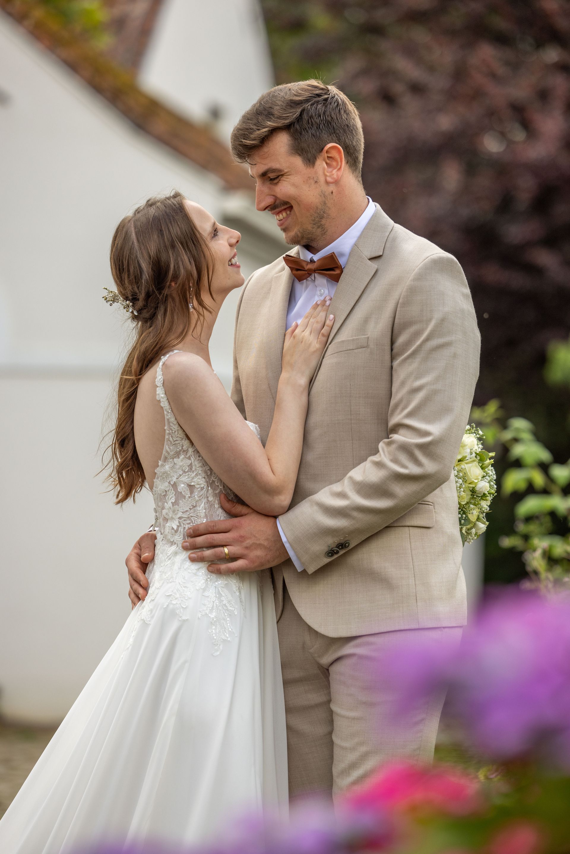 Bruid en bruidegom omhelzen elkaar en glimlachen. Hij draagt ​​een bruin pak en vlinderdas, zij een witte trouwjurk. Buiten.