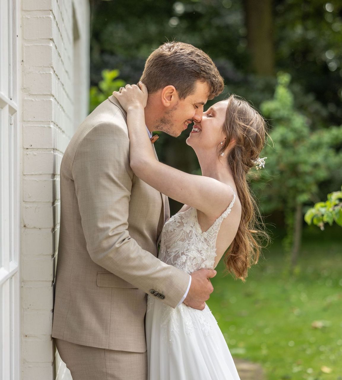 Bruid en bruidegom omhelzen elkaar en staan ​​op het punt elkaar te kussen. Ze staan ​​voor een wit gebouw, omringd door groen.