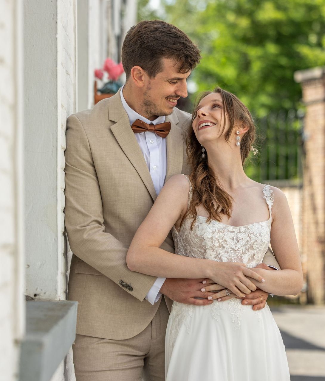 Een man in een bruin pak met een bruine vlinderdas omhelst een vrouw in een trouwjurk. Ze lachen naar elkaar.