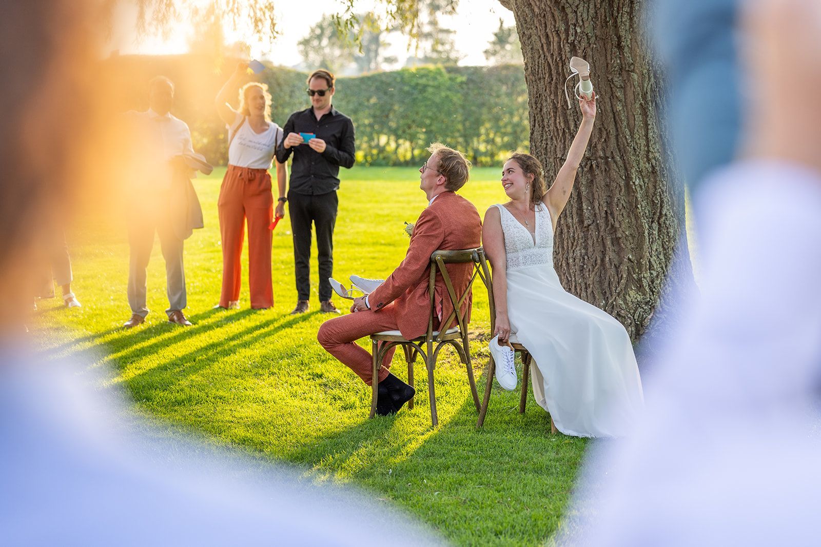 De bruid tilt haar schoenen op, de bruidegom zit op haar stoel, de gasten kijken buiten toe op het zonnige gazon.