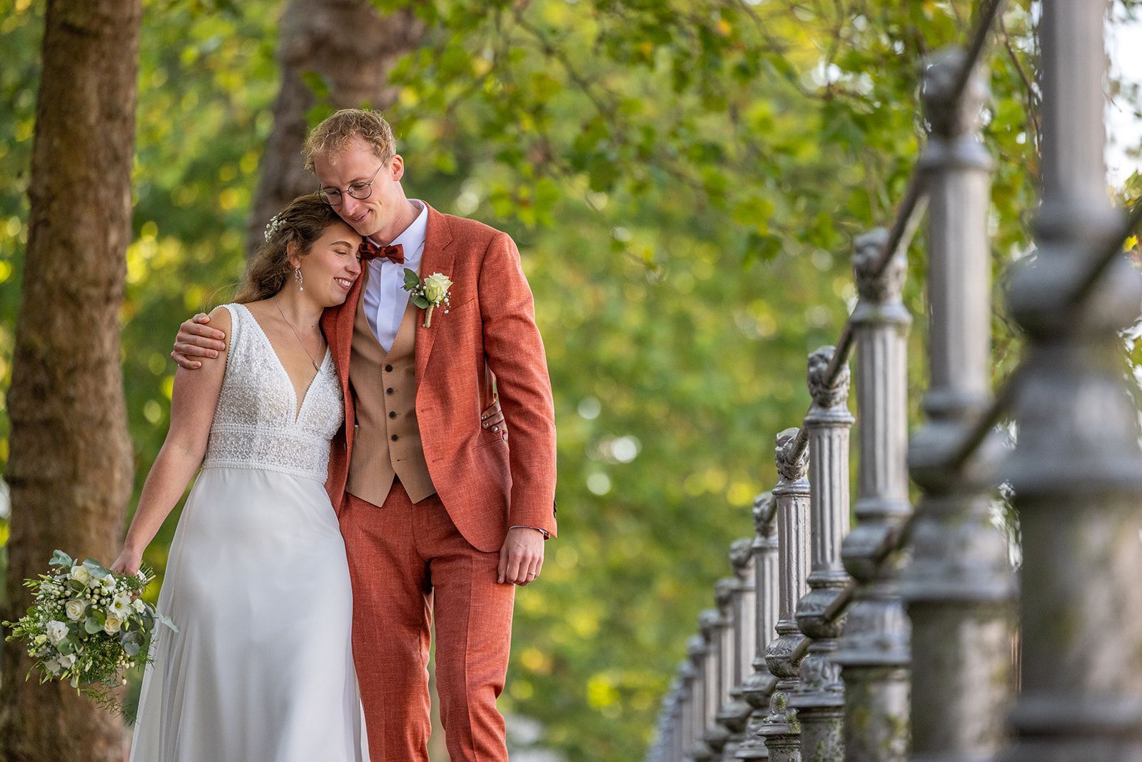 Het pasgetrouwde stel omarmt de buitenlucht. Bruid in witte jurk, bruidegom in roestbruin pak, langs een reling.
