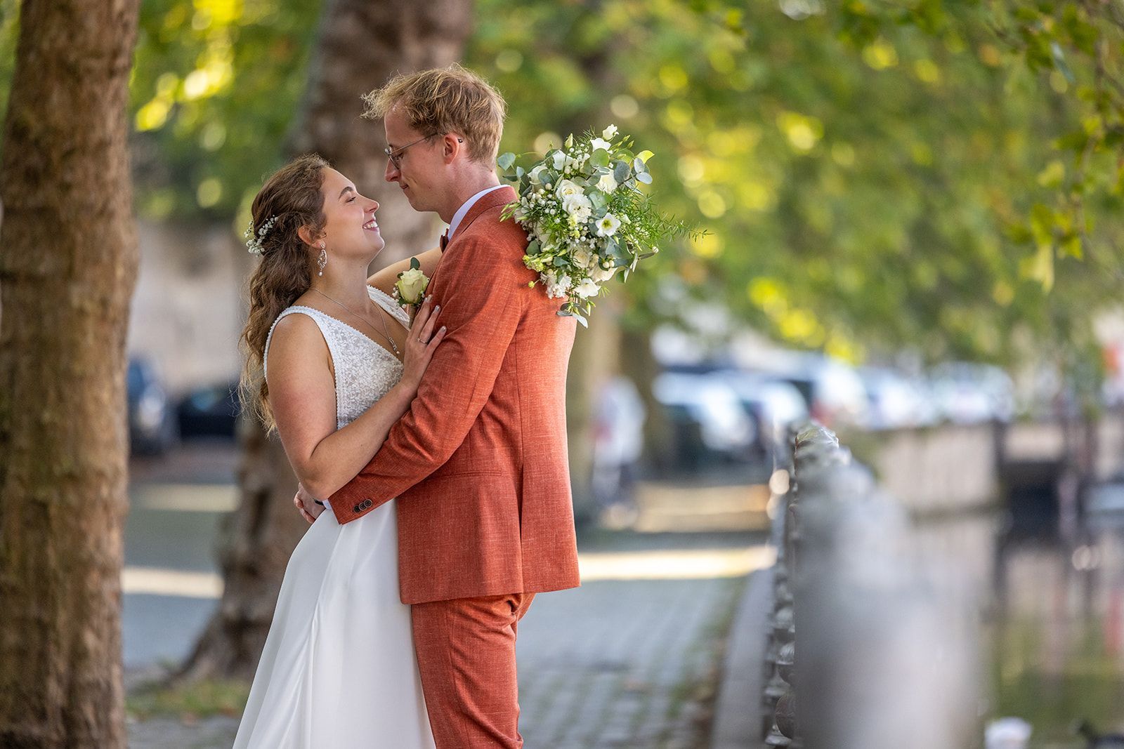 Bruidspaar omhelst elkaar buiten, bruidegom in oranje pak met boeket, bruid in witte jurk lachend.