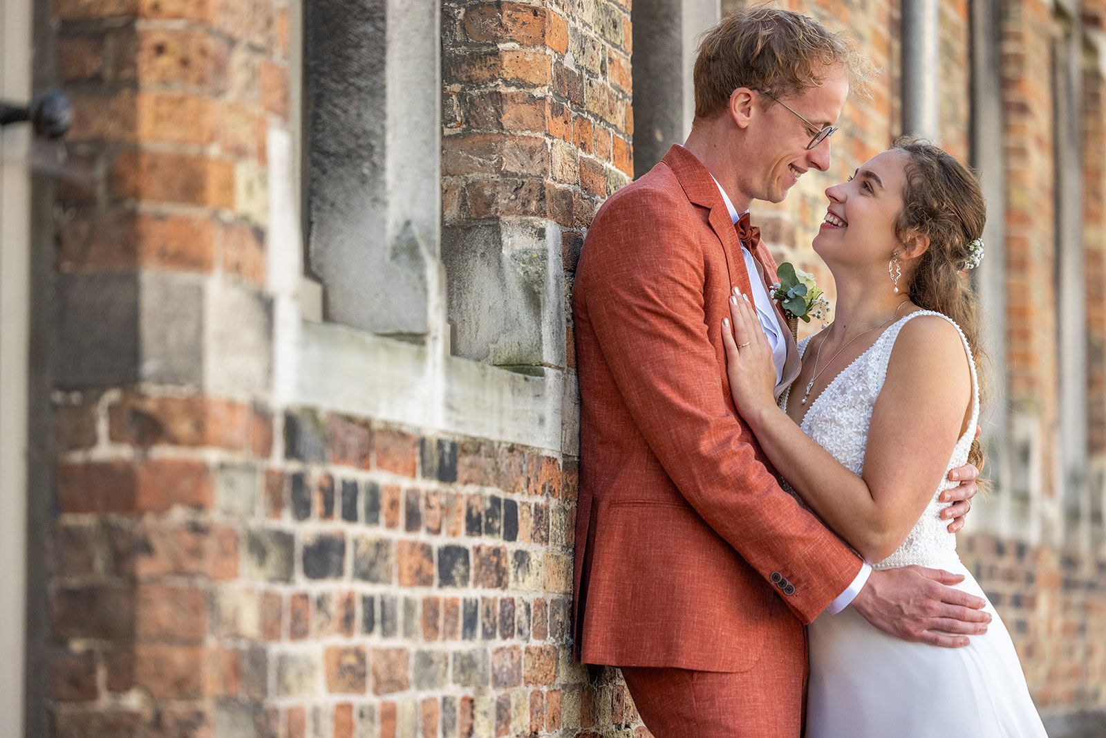 Bruid en bruidegom omhelzen elkaar tegen een bakstenen gebouw en glimlachen naar elkaar. De bruidegom draagt ​​een oranje pak.