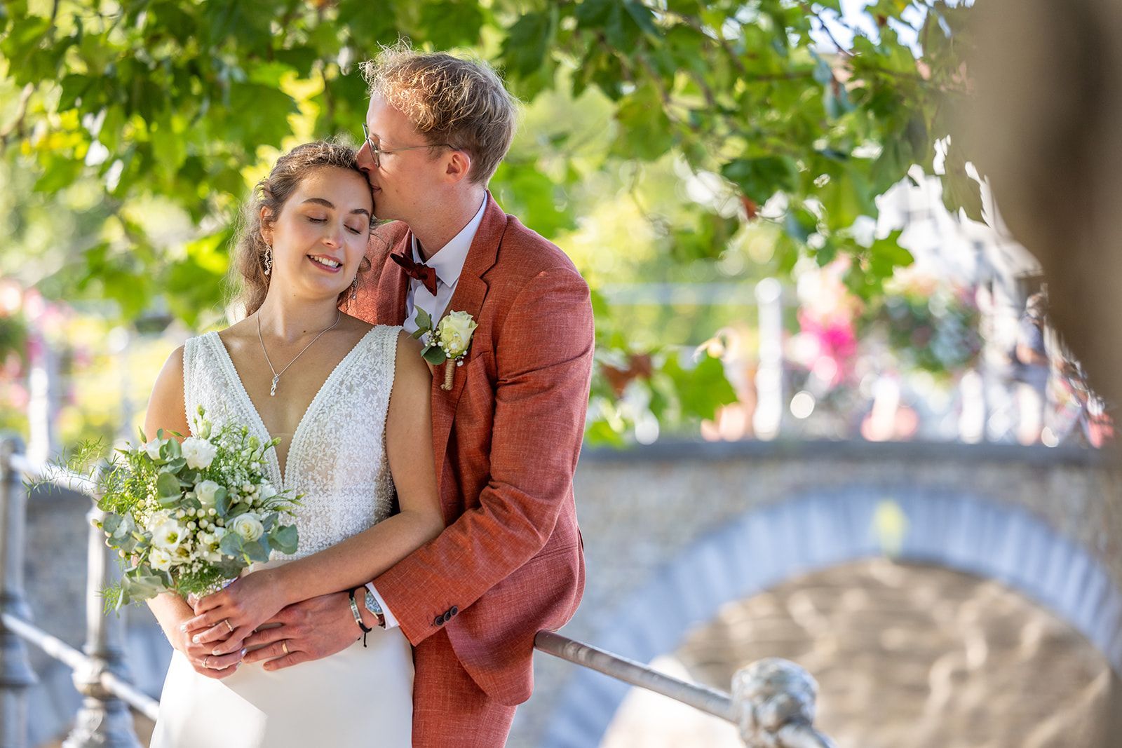 Pasgetrouwden omhelzen elkaar bij een stenen brug. De man kust de vrouw op het voorhoofd; zij houdt bloemen vast. Zonnig, buiten.