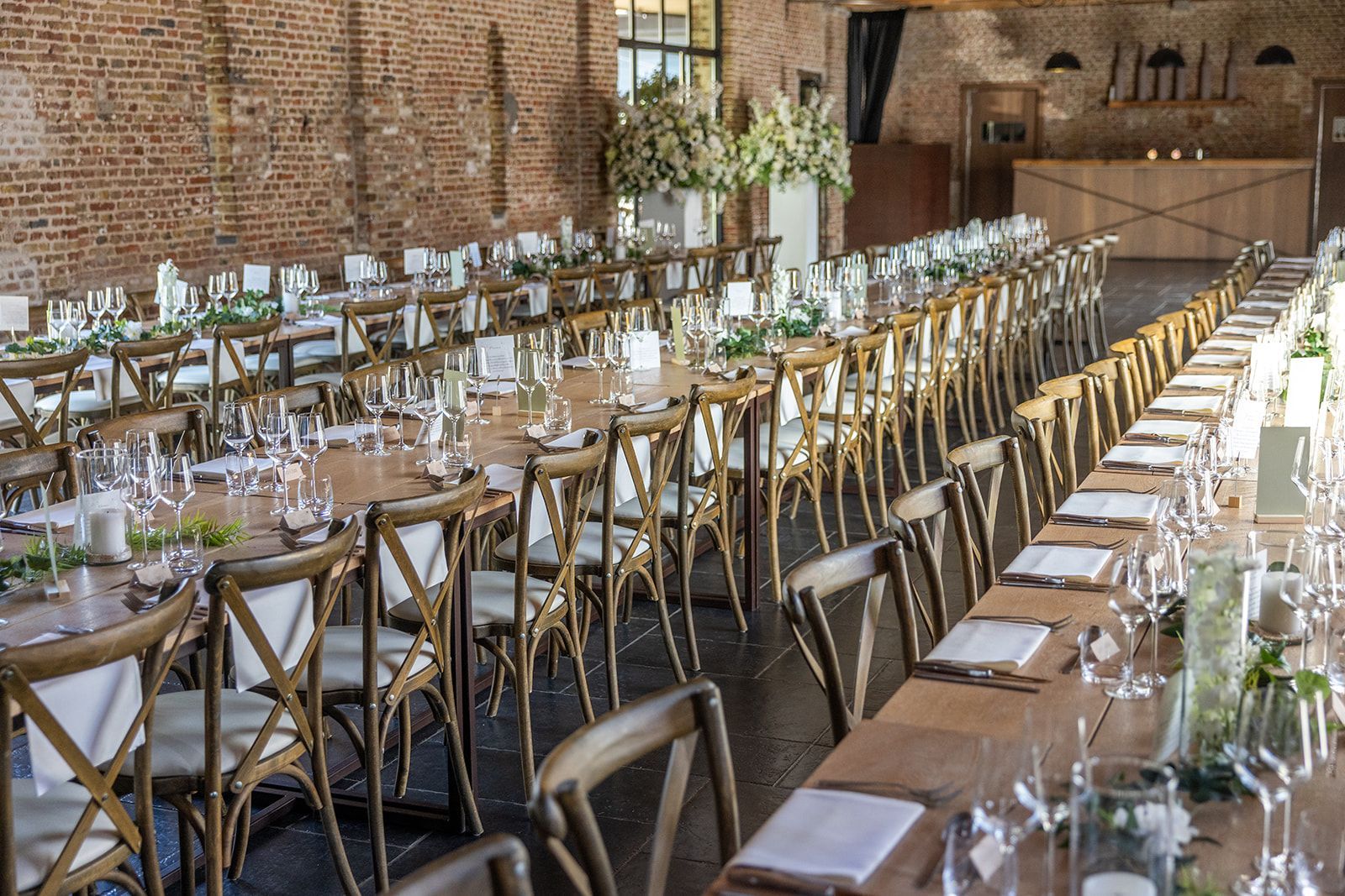 Lange, feestelijk gedekte tafels voor een formeel evenement in een ruimte met bakstenen muren, houten stoelen en bloemstukken.