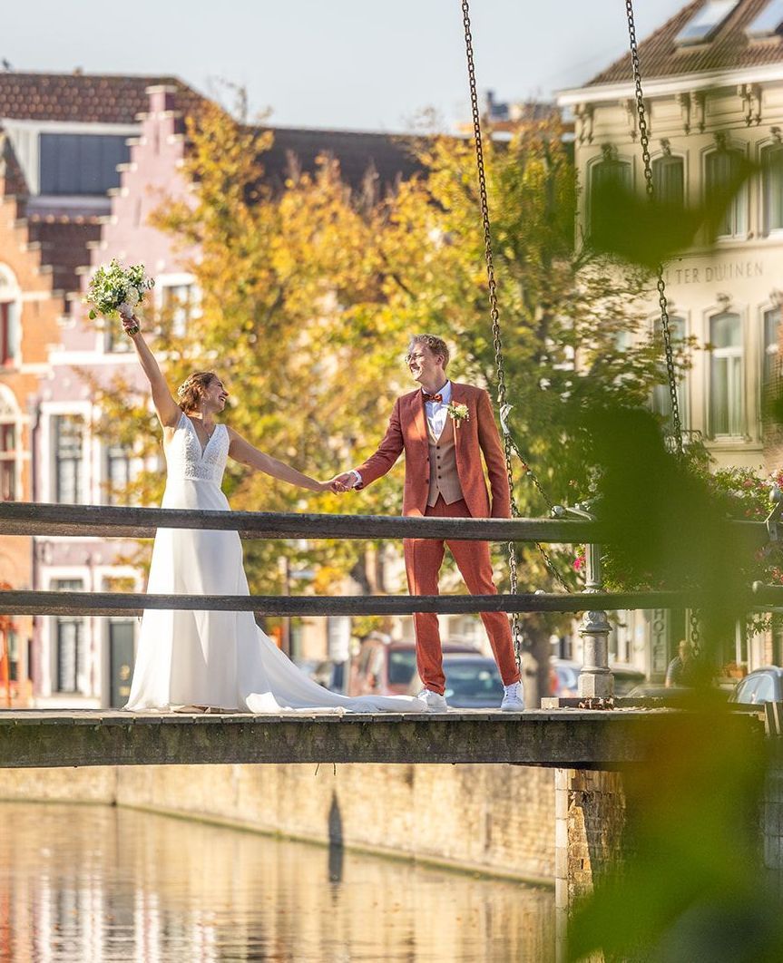 Pasgetrouwden op een brug, hand in hand en lachend. De vrouw in een witte jurk, de man in een roestbruin pak. Gebouwen op de achtergrond.