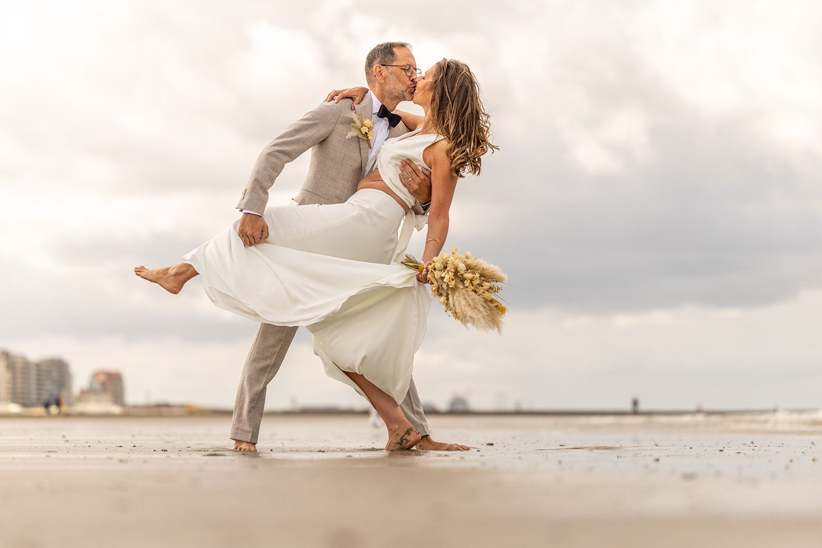 Bride and groom dip kissing on beach, bride's skirt billowing, groom in suit, cloudy sky.