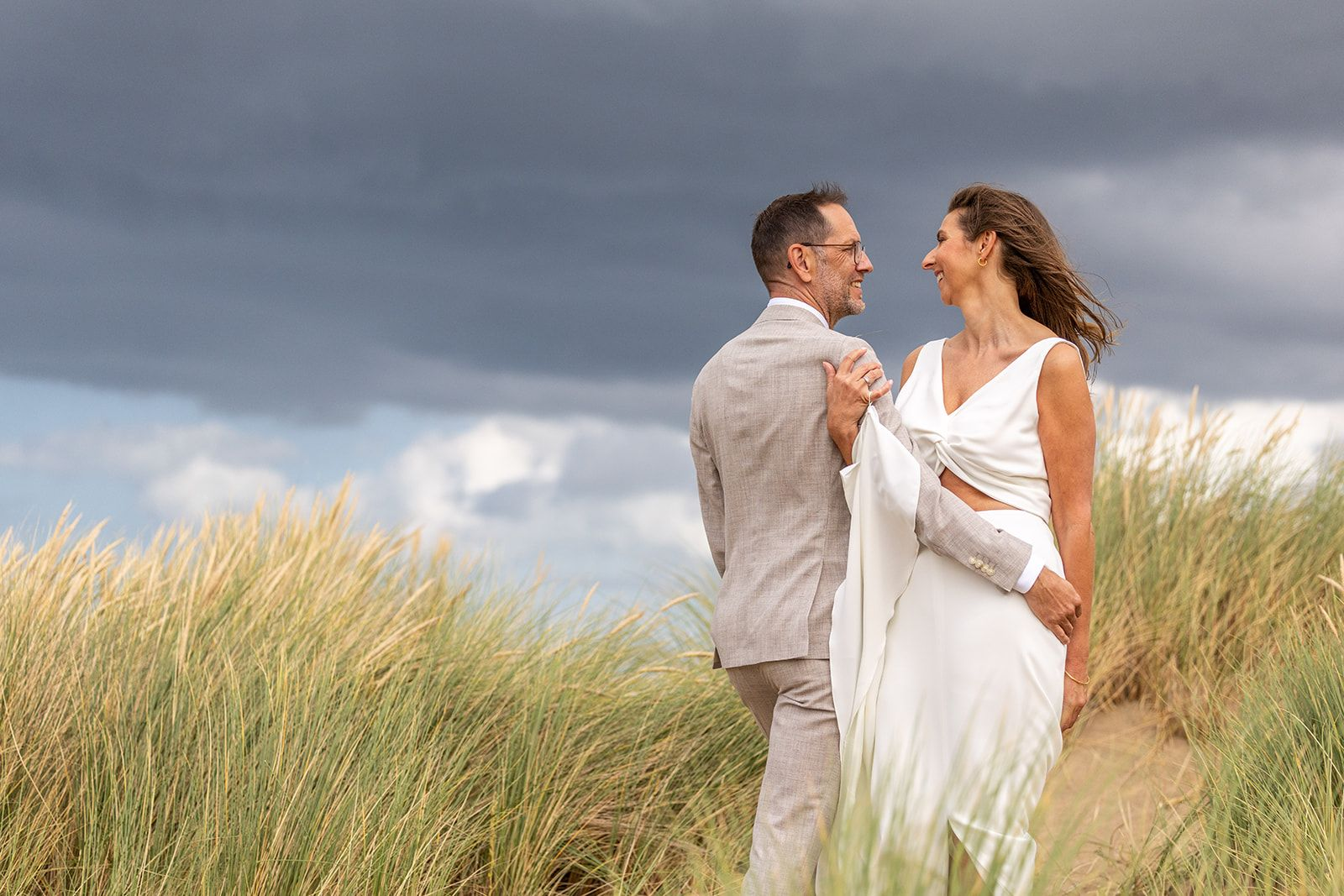 Bruid en bruidegom hand in hand, wandelend op het strand; gebouwen op de achtergrond.