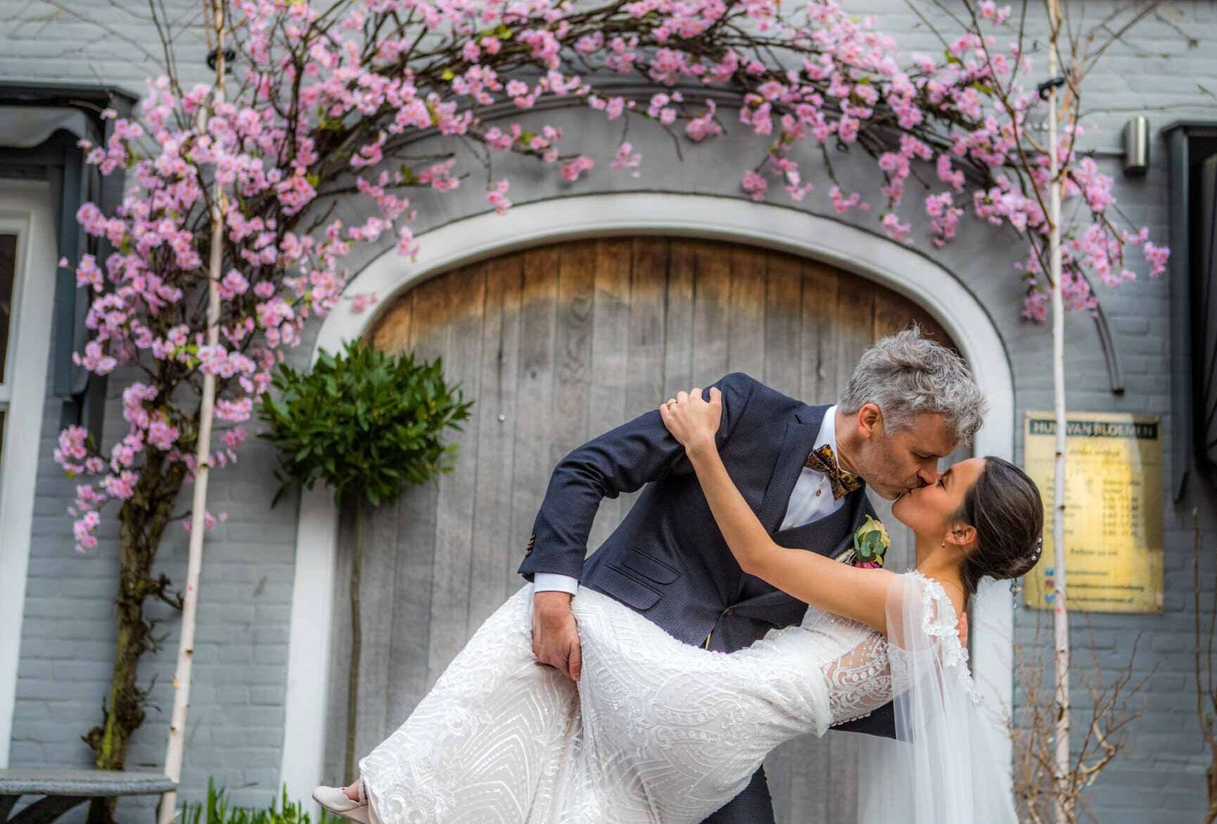 Kussend paar onder een boog met roze bloemen; bruid in witte jurk, bruidegom in pak.