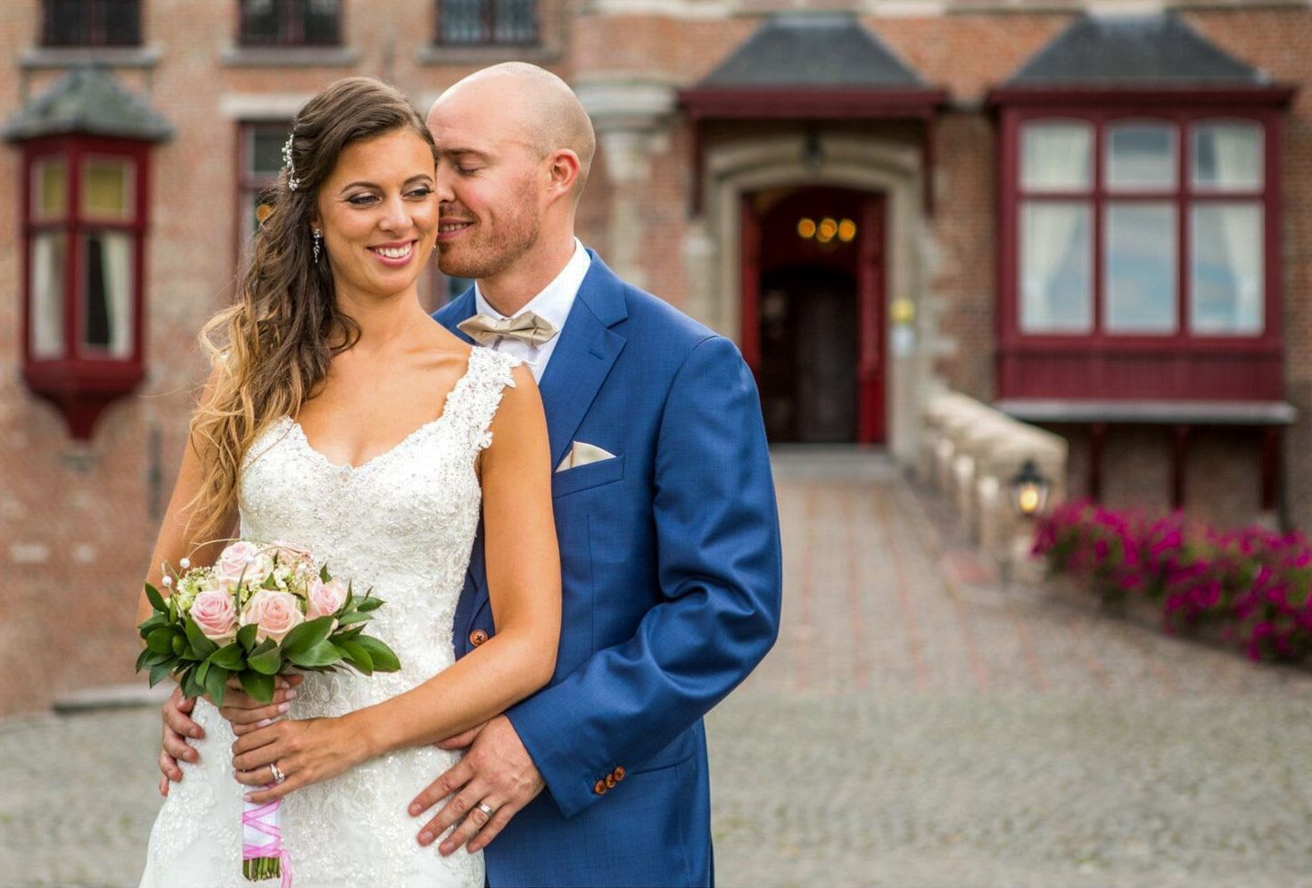 Bruid en bruidegom omhelzen elkaar voor een bakstenen gebouw. ​​De bruid in de witte jurk houdt bloemen vast, de bruidegom in het blauwe pak.
