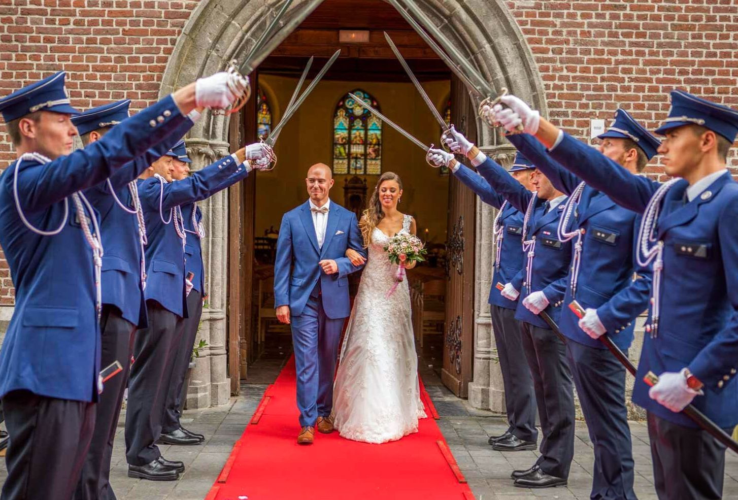 Bruid en bruidegom verlaten de kerk onder zwaarden vastgehouden door mensen in uniform op de rode loper.