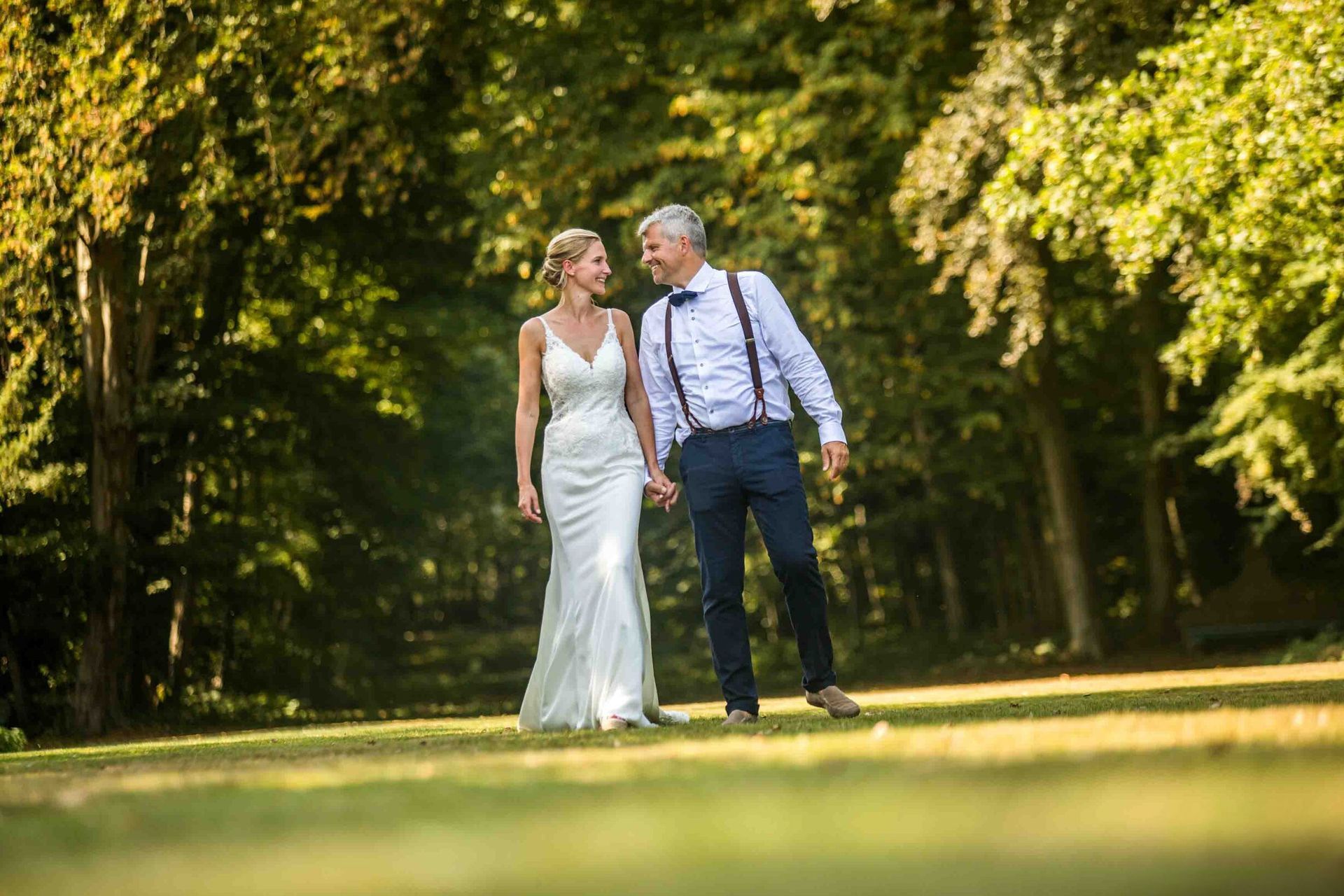 Een stel houdt elkaars hand vast en lacht naar elkaar terwijl ze over een graspad in een zonnig park lopen.