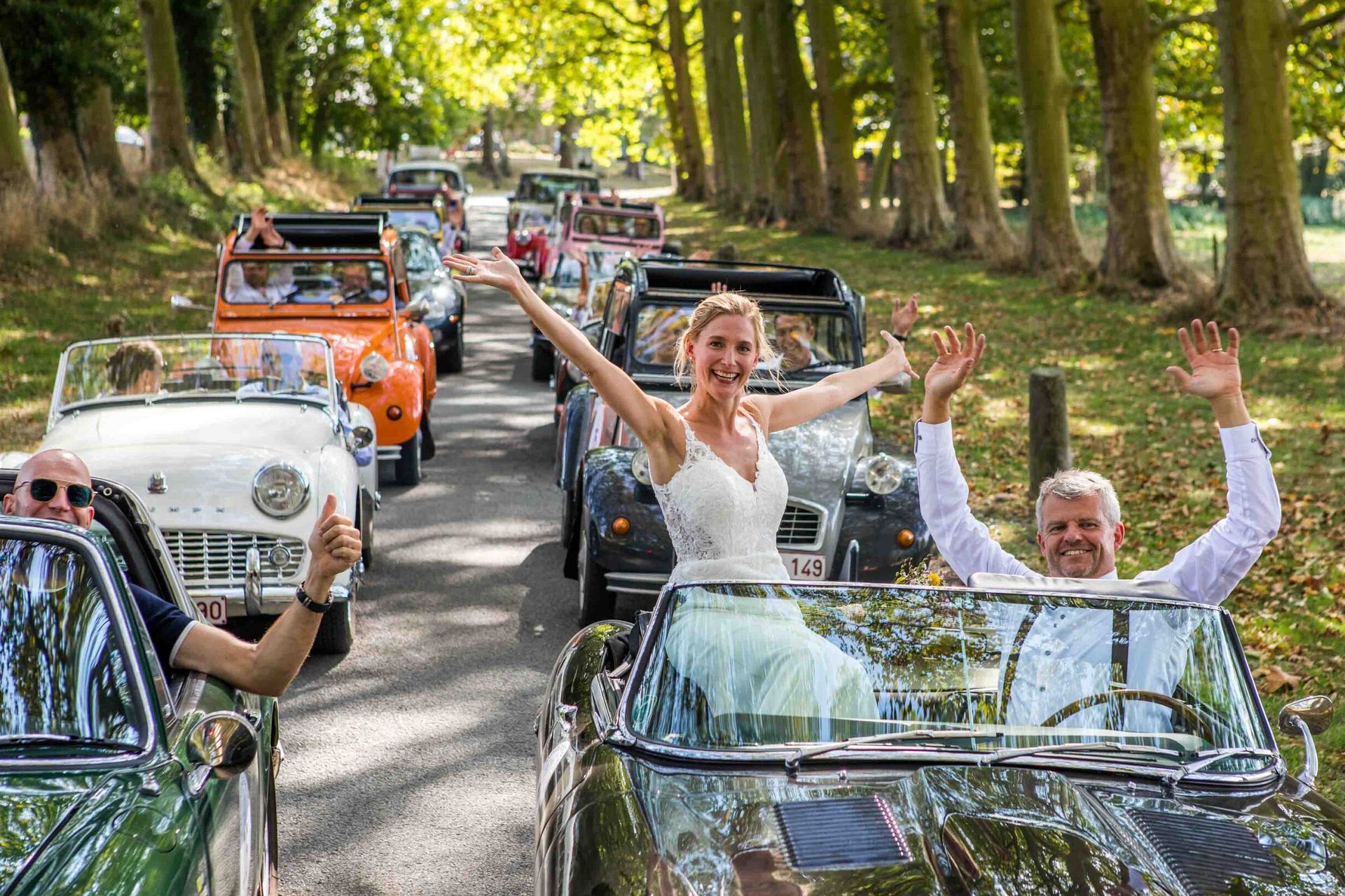Bruiloftsstoet in klassieke auto's, bruid en bruidegom zwaaien, opgesteld op een met bomen omzoomde weg.