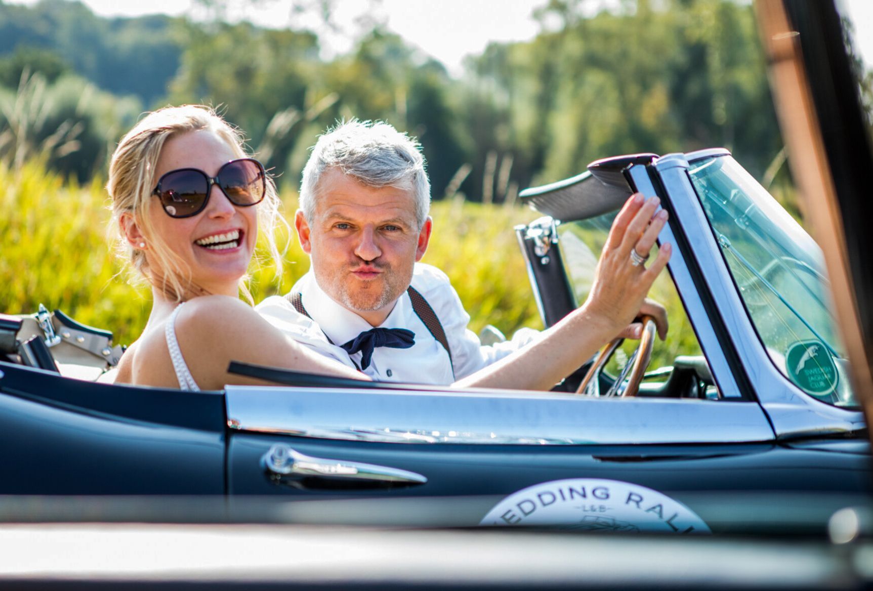 Pasgetrouwd stel in een klassieke cabriolet, lachende vrouw, man die een raar gezicht trekt, zonnig plekje buiten.